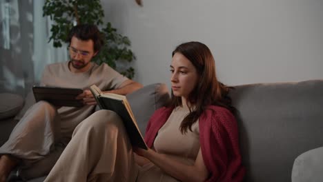 Close-up-of-a-happy-young-brunette-girl-in-a-red-sweater-sitting-on-a-gray-sofa-with-her-boyfriend-and-reading-a-book-while-a-brunette-man-is-looking-at-something-on-a-black-tablet-in-a-modern-apartment