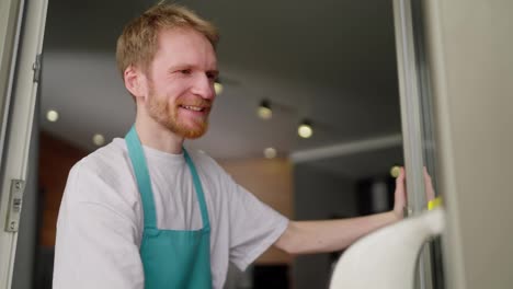 Side-view-of-a-confident-blond-guy-in-a-white-T-shirt-and-blue-apron-washes-a-glass-door-using-a-window-vacuum-cleaner-during-a-gentle-cleaning-of-the-apartment