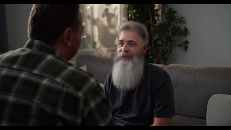 Over-the-shoulder-A-happy-and-joyful-elderly-man-with-gray-hair-and-a-lush-gray-beard-with-an-earring-in-his-ear-and-in-a-gray-T-shirt-shakes-hands-with-his-boyfriend-with-black-hair-in-a-checkered-shirt-and-shows-his-emotions-and-feelings-while-sitting-on-a-gray-sofa-in-a-modern-apartment-in-the-ev