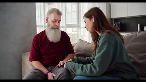 Un-Hombre-Mayor-De-Pelo-Gris-Y-Barba-Frondosa-Con-Una-Camiseta-Roja,-Junto-Con-Su-Hija-Morena-Adulta-Con-Una-Chaqueta-Verde,-Miden-Su-Pulso-Usando-Un-Dispositivo-Conectado-A-Su-Dedo-Mientras-Están-Sentados-En-El-Sofá-De-Un-Apartamento-Moderno.