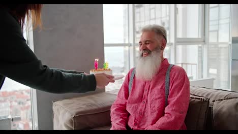 A-happy-man-with-gray-hair-of-advanced-age-with-a-lush-beard-in-a-pink-shirt-receives-a-cake-and-candles-from-his-adult-daughter-in-a-green-jacket-in-a-modern-apartment