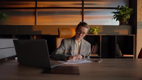 Confident-middle-aged-business-woman-with-glasses-in-a-gray-jacket-and-business-uniform-sits-at-a-wooden-table-and-writes-down-her-ideas-in-front-of-a-laptop-in-a-sunny-office