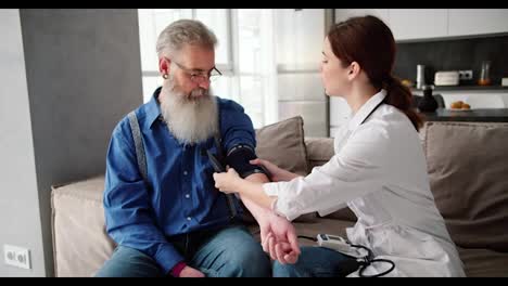 A-brunette-girl-in-a-white-coat-the-doctor-expresses-her-verdict-after-an-examination-with-a-heart-rate-monitor-and-a-blood-pressure-examination-of-an-elderly-man-with-gray-hair-and-a-lush-beard-in-a-blue-shirt-on-the-sofa-at-home