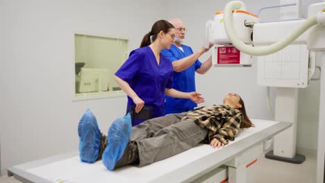 A-young-brunette-girl-in-a-checkered-shirt-lies-on-a-bed-in-an-x-ray-room-while-experienced-doctors-an-elderly-man-and-a-girl-in-a-blue-uniform-perform-x-rays-to-diagnose-a-young-girl-and-prevent-diseases-in-a-modern-clinic