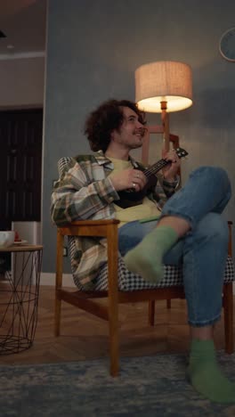 Vertical-video-of-a-happy-brunette-guy-with-curly-hair-and-a-mustache-in-a-checkered-shirt-sitting-on-a-chair-and-playing-the-ukulele-at-home-near-the-lamp