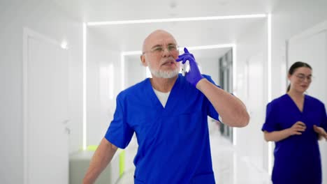 Un-Médico-Anciano,-Con-Gafas-Y-Barba-Blanca-Y-Un-Uniforme-Azul,-Corre-A-Toda-Prisa-Por-El-Pasillo-Hablando-Con-Un-Teléfono-Inteligente-En-Una-Clínica-Moderna