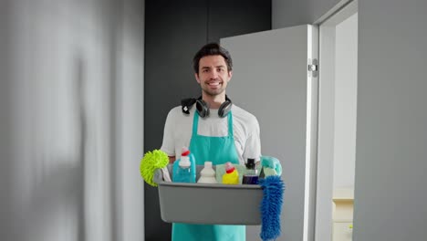 Zoom-in-confident-guy-brunette-cleaner-in-a-white-T-shirt,-blue-apron-and-black-wireless-headphones-holds-a-gray-plastic-basin-with-cleaning-tools-for-cleaning-and-detergents-in-a-modern-bright-apartment