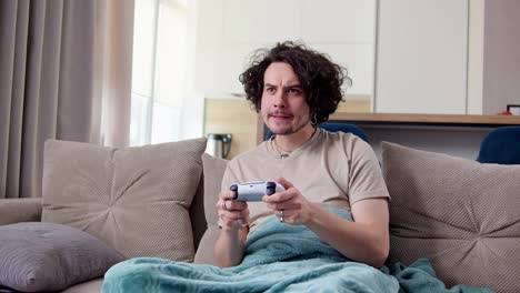 Happy-brunette-guy-with-curly-hair-and-mustache-plays-video-games-using-a-white-joystick-while-sitting-on-a-sofa-with-a-blue-blanket-in-a-modern-apartment