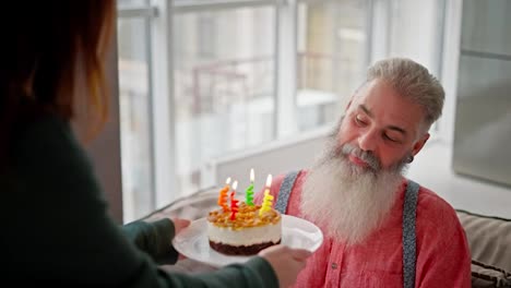 A-happy-man-with-gray-hair-and-a-lush-beard-in-a-pink-shirt-blows-out-four-candles-and-on-a-small-cake-during-his-birthday-and-congratulations-from-his-adult-daughter-in-a-modern-apartment