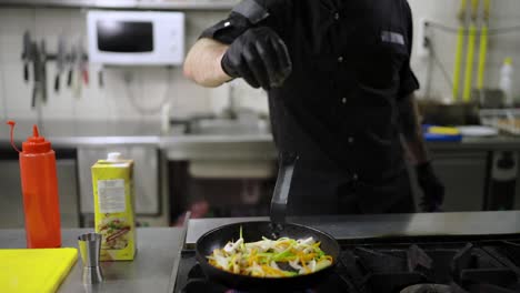 Primer-Plano-De-Un-Chef-Confiado-Con-Guantes-Negros-Rocía-Verduras-Con-Condimentos-Y-Luego-Las-Mezcla-Eficazmente-En-Una-Estufa-De-Gas-En-La-Cocina-De-Un-Restaurante.-Un-Chef-Confiado-Sacude-Una-Sartén-Para-Revolver-Las-Verduras-Que-Se-Fríen-En-Una-Sartén.