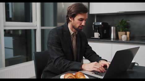 A-serious-and-confident-brunette-man-in-a-black-jacket-and-shorts-works-at-a-gray-laptop-while-sitting-at-the-dining-table-in-the-kitchen-working-remotely-in-a-modern-apartment