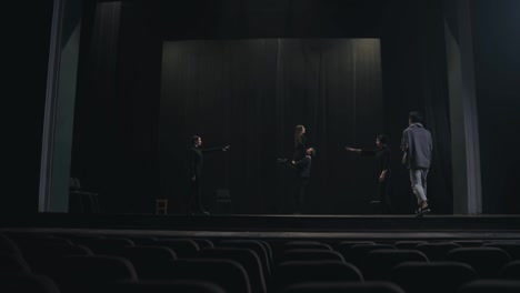 Vista-Desde-La-Audiencia:-Dos-Actores-Confiados-Interpretan-La-Escena-Final-Con-Su-Director-De-Escena-Con-Una-Camiseta-Gris-Y-Una-Bufanda-Durante-Su-Ensayo-En-El-Escenario-En-Un-Teatro-Vacío-Con-Cortinas-Negras.