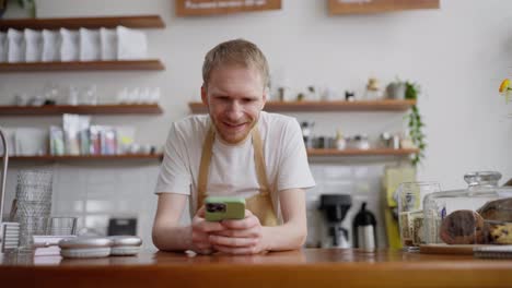 Happy-blond-guy-barista-in-a-yellow-apron-stands-behind-the-counter-and-types-on-social-networks-in-a-cafe