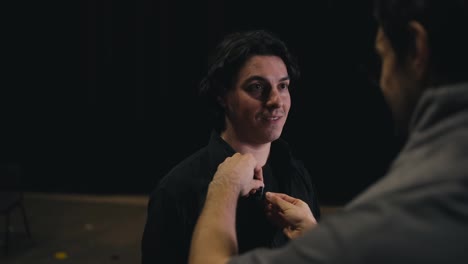 Over-the-shoulder-a-confident-male-stage-director-in-gray-clothes-attaches-a-small-microphone-to-a-young-brunette-actor-in-a-black-suit-before-going-on-stage-in-a-theater-during-a-performance