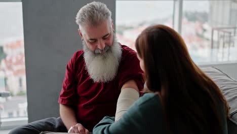 Un-Anciano-Feliz-Con-Cabello-Gris-Y-Una-Barba-Exuberante-Con-Una-Camiseta-Roja-Ayuda-A-Vendar-Su-Mano-A-Su-Hija-Adulta-Morena-Con-Un-Suéter-Verde-En-Un-Apartamento-Moderno.