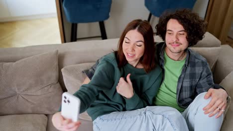 Ein-Glückliches-Brünettes-Mädchen-Macht-Ein-Selfie-Mit-Einem-Weißen-Smartphone-Mit-Ihrem-Brünetten-Freund-Mit-Lockigem-Haar-Zu-Hause-Auf-Dem-Sofa-In-Einem-Modernen-Apartment