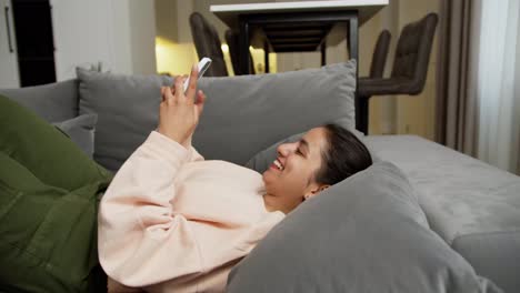 A-happy-brunette-girl-with-Brown-skin-in-a-light-sweater-and-green-pants-lies-on-a-gray-sofa-and-types-on-her-phone-happy-chatting-with-her-friends-online-at-home