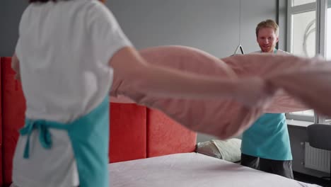 Confident-group-of-two-cleaners-a-blond-guy-and-a-brunette-girl-in-a-white-T-shirt-and-blue-aprons-make-the-bed-with-a-pink-blanket-during-cleaning-in-the-bedroom