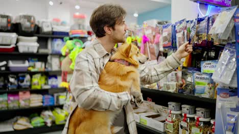 Confident-brunette-guy-holding-his-red-corgi-in-his-arms-while-shopping-and-inspecting-goods-in-a-pet-store