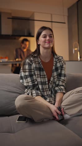 Vertical-video-of-a-happy-brunette-girl-in-a-plaid-shirt-sitting-on-a-gray-sofa-and-switching-channels-on-the-TV-using-the-remote-control-in-a-modern-apartment-at-home-near-her-boyfriend-in-the-background-who-is-working-on-his-laptop