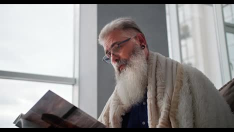 An-elderly-man-with-gray-hair-and-a-lush-well-groomed-beard-is-wrapped-in-a-white-blanket-and-reading-a-magazine-while-sitting-on-the-sofa-in-a-modern-apartment