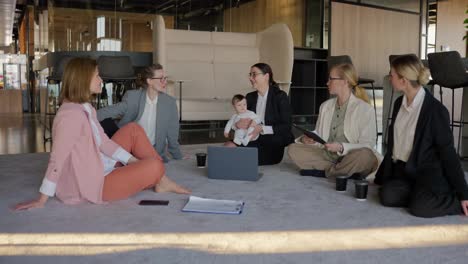 A-group-of-businesswoman-in-business-clothes-sit-in-a-circle-on-a-gray-carpet-and-communicate-while-a-brunette-girl-in-a-business-suit-holds-her-little-baby-daughter-in-her-arms-in-a-modern-office