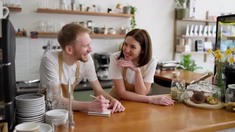 Happy-brunette-girl-in-a-yellow-apron-and-white-T-shirt-communicates-with-her-colleague-and-laughs-while-writing-important-thoughts-and-ideas-in-a-notebook-in-a-cafe