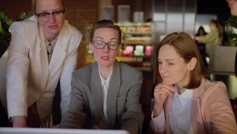 A-group-of-confident-businesswomen-in-a-business-uniform-gathered-around-a-laptop-and-a-girl-speaker-in-a-gray-suit-presenting-their-work-and-ideas-while-working-in-a-modern-office