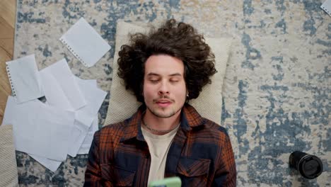 A-tired-brunette-guy-with-curly-hair-and-a-mustache-lies-on-the-floor-and-tries-to-rest-on-a-pillow-at-home-in-a-modern-apartment