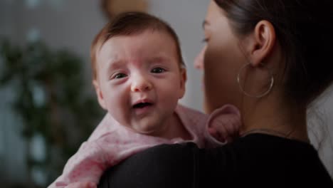 Close-up-portrait-of-a-little-baby-girl-in-a-pink-suit-sitting-in-her-mother-arms-and-looking-over-her-shoulder-posing-in-a-modern-apartment