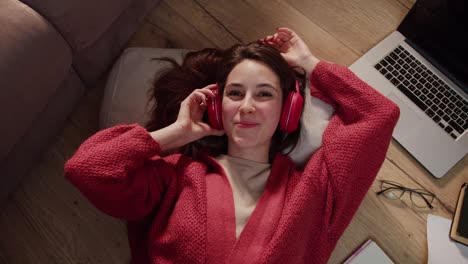 Portrait-of-a-confident-and-happy-young-brunette-girl-in-a-red-sweater-and-wireless-headphones-who-listens-to-music-during-her-break-between-work-in-a-modern-apartment-on-the-floor