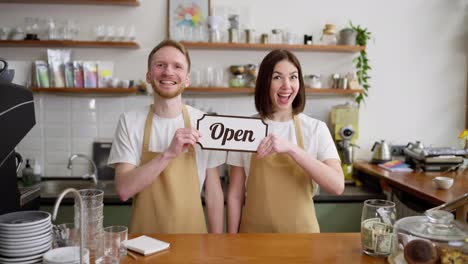 Portrait-of-a-happy-brunette-girl-in-a-yellow-apron-together-with-her-colleague-a-blond-guy-raising-a-sign-with-an-open-sign-while-working-in-a-cafe