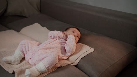 Side-view-of-a-little-girl-baby-in-a-pink-jumpsuit-lies-on-a-special-mat-on-the-sofa-in-a-modern-apartment.-Close-up-shot-of-a-little-girl-baby-in-a-pink-overalls-and-white-socks-lying-on-a-special-bedding-on-pillows-on-the-sofa