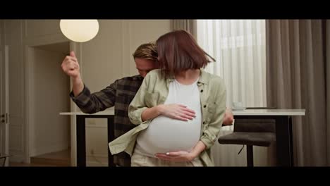 Happy-blond-man-in-a-plaid-shirt-and-white-T-shirt-dances-with-his-pregnant-brunette-wife-in-a-white-T-shirt-in-a-modern-apartment-during-the-day.-Happy-man-dancing-freestyle-with-his-pregnant-brunette-girlfriend