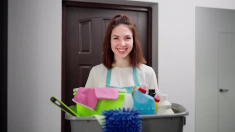 Portrait-of-a-happy-confident-brunette-girl-and-a-cleaning-lady-in-a-blue-apron-with-a-gray-plastic-basin-in-her-hands-in-which-there-are-a-large-number-of-different-tools-for-cleaning-a-modern-apartment-on-call