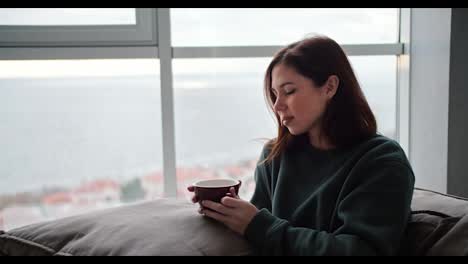 A-brunette-girl-in-a-green-jacket-sits-on-the-sofa-holds-a-pillow-on-her-legs-and-drinks-hot-tea-from-a-brown-mug-in-a-modern-apartment-overlooking-the-sea