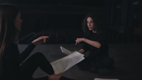 A-confident-and-interested-brunette-girl-with-curly-hair-in-round-glasses-and-a-black-theatrical-costume-communicates-with-her-colleague-and-they-play-roles-preparing-to-study-for-a-performance-on-stage-in-an-empty-theater