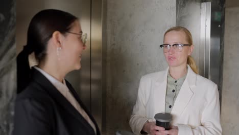 Confident-brunette-girl-in-round-glasses-in-a-business-uniform-talking-with-her-colleague-blonde-businesswoman-while-they-are-drinking-coffee-during-a-break-between-work-in-a-modern-office