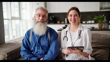 Portrait-of-an-elderly-man-with-a-lush-gray-beard-and-glasses-in-a-blue-shirt-who-sits-on-the-sofa-with-a-female-doctor-in-a-white-coat-who-holds-a-tablet-during-a-home-examination-in-a-modern-apartment