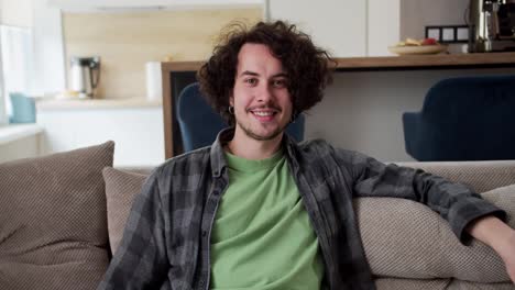 Portrait-of-a-happy-brunette-guy-with-curly-hair-a-mustache-and-a-plaid-shirt-who-sits-on-the-sofa-in-a-modern-studio-apartment