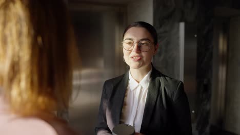 Over-the-shoulder-a-confident-brunette-girl-in-round-glasses-and-a-black-business-uniform-communicates-with-her-colleague-while-drinking-coffee-in-a-modern-sunny-office-during-work