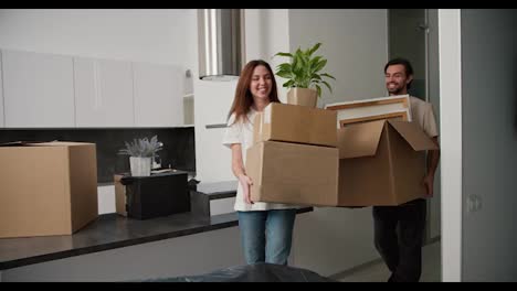 A-happy-brunette-girl-in-a-white-T-shirt-and-jeans-together-with-her-happy-brunette-boyfriend-with-stubble-walk-into-their-new-apartment-with-boxes-in-their-hands-and-put-them-on-a-closed-sofa-in-a-new-studio-apartment-modern