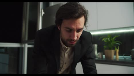 Serious-brunette-man-with-stubble-wipes-sweat-from-his-forehead-in-a-black-jacket-and-beige-shirt-working-in-front-of-his-laptop-and-then-rejoicing-at-his-success-moving-away-from-his-desk-showing-his-beige-shorts-in-a-modern-apartment-in-the-evening