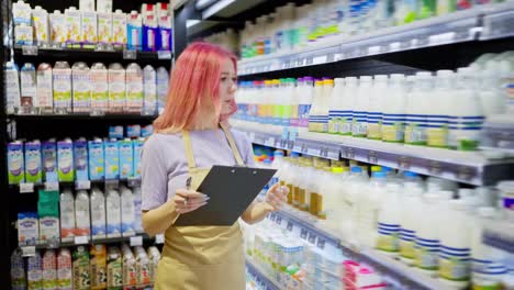 A-girl-supermarket-worker-with-pink-hair-holds-a-tablet-in-her-hands-and-takes-inventory-of-dairy-products-on-a-shelf-in-a-supermarket