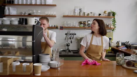 Confident-brunette-waitress-cleans-the-counter-with-a-pink-rag-and-communicates-with-a-blond-guy-at-the-beginning-of-the-working-day-in-a-cafe