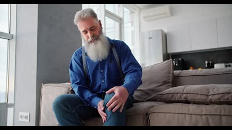 Ein-älterer-Mann-Mit-Grauem-Haar-Und-üppigem-Bart-In-Einem-Blauen-Hemd-Streichelt-Und-Massiert-Sein-Knie,-Während-Er-Auf-Einem-Braunen-Modernen-Sofa-In-Einer-Wohnung-Sitzt