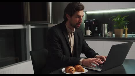 A-serious-brunette-man-with-stubble-in-a-black-suit-sits-at-a-table-in-front-of-a-laptop-and-communicates-via-video-conference-while-sitting-without-pants-only-in-shorts-in-a-modern-kitchen-in-the-evening-at-home