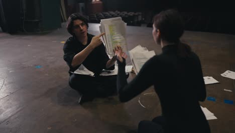 Two-confident-actors-a-brunette-girl-and-a-man-in-a-black-suit-are-sitting-on-stage-sorting-through-sheets-of-paper-with-a-script-talking-about-their-role-and-rehearsals-for-the-play