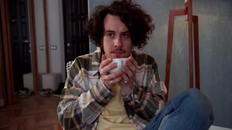 Anxious-brunette-guy-with-curly-hair-in-a-checkered-shirt-drinks-tea-from-a-white-cup-brings-the-cup-with-both-hands-to-his-lips-and-thinks-in-a-modern-apartment