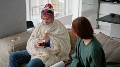 Un-Hombre-Mayor-Con-Un-Sombrero-Rojo-Y-Una-Manta-Blanca-Bebe-Té-Caliente-De-Una-Taza-Marrón-Y-Se-Comunica-Con-Su-Hija-Morena-Adulta-Con-Una-Chaqueta-Verde-Mientras-Está-Sentado-En-El-Sofá-De-Un-Apartamento-Moderno.